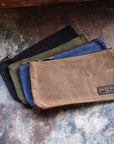 Waxed canvas zip pouches in black, olive, blue and tan fanned out on a weathered metal surface