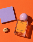 Dolly perfume bottle beside a pistachio and a lavender block on an orange background