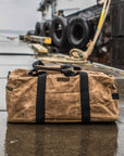 Tan waxed canvas duffel bag on a wet dock with tires and rope in the background