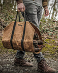 Person in jeans and boots carrying the tan waxed canvas firewood carrier full of logs through the woods