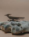 A dark metal bird silhouette candle holder mounted on a textured gray stone base, photographed against a solid warm beige background.
