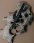 A black metal cactus bottle opener mounted on a textured, organic-shaped gray stone base, set against a solid warm brown background.