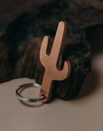A cactus-shaped keychain made of copper-colored metal with a silver split key ring, resting against a dark textured wood prop on a beige background.
