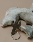 A cactus-shaped metal keychain tool with a silver split ring, resting against a light gray decorative bone on a warm beige background.