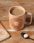 Ceramic mug with sun design on a wooden surface, next to a notebook and spoon with sugar.