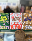 Three 'Date Better' chocolate packaging boxes on a glass shelf with blurred background