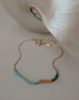 A delicate gold chain bracelet with small turquoise and amber beads, resting on a textured light beige surface next to a white shell.