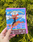 Hand holding a colorful birthday card with a horse and rider design against a green leafy background.