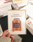 Card with a humorous design of a jar of cheese balls and text, held in front of a stack of papers.