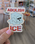 Sticker with 'ABOLISH ICE' text held in a hand against a blurred library background