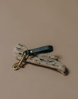 A rustic keychain or bag charm featuring a dark navy leather loop attached to a weathered, speckled off-white driftwood piece with a gold-tone metal swivel clasp, set against a solid warm beige background.