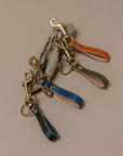 Four leather strap keychains in blue, orange, green, and gray, each attached to a gold-tone swivel clasp and displayed on a neutral beige background.
