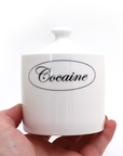 White container with 'Cocaine' label held by a hand on a white background