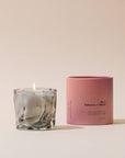 Smoky gray wavy glass candle beside a pink cylindrical box on a cream backdrop