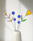Blue thistles, pale leaf stems and an orange glass flower in a white ribbed vase in soft window light
