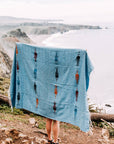 Person holding up a light blue thunderbird blanket on a cliff above the ocean