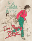 A close-up of a beige fabric surface printed with a retro-style illustration and the text 'Not tonight Honey, I'm Playing Bingo.' The print shows a woman in a pink top and green pants walking away from a bed, with a man in bed, a cat, and bingo cards.