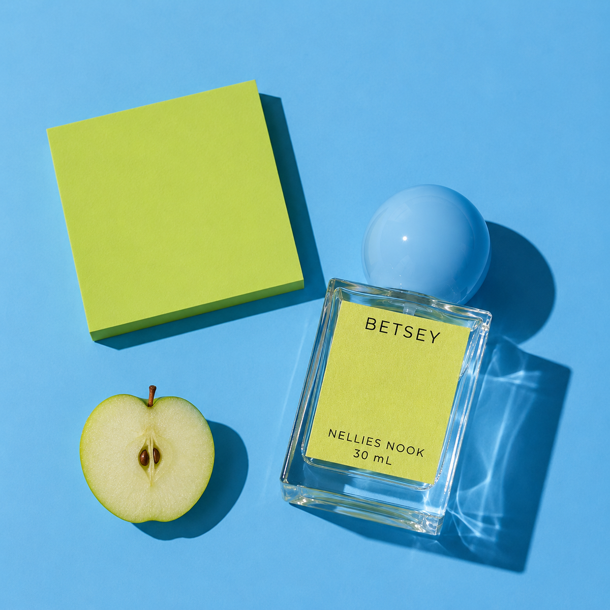 Betsey perfume bottle beside half a green apple and a chartreuse block on a blue background