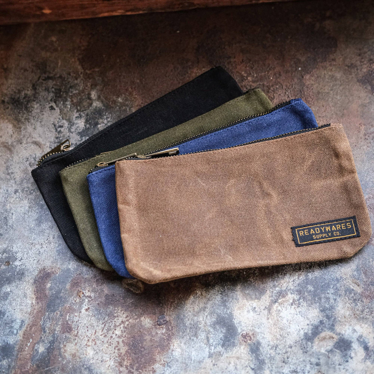 Waxed canvas zip pouches in black, olive, blue and tan fanned out on a weathered metal surface