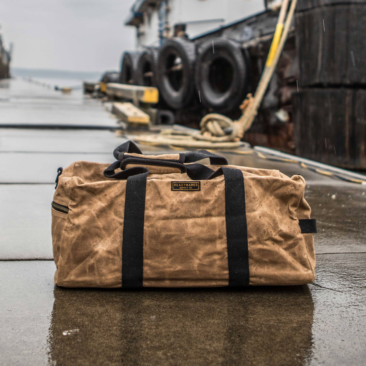 Tan waxed canvas duffel bag on a wet dock with tires and rope in the background