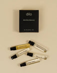 Five small perfume sample vials with black spray caps are arranged on a warm beige background below a black rectangular box labeled 'dilo elsewhere discovery eau de parfum / 5 ml'. The vials contain clear, yellow, and white liquids, with some labels partially visible including 'Sakura Oud', 'Citrus Flower', 'Desert Kush', and 'Pale Samso'.