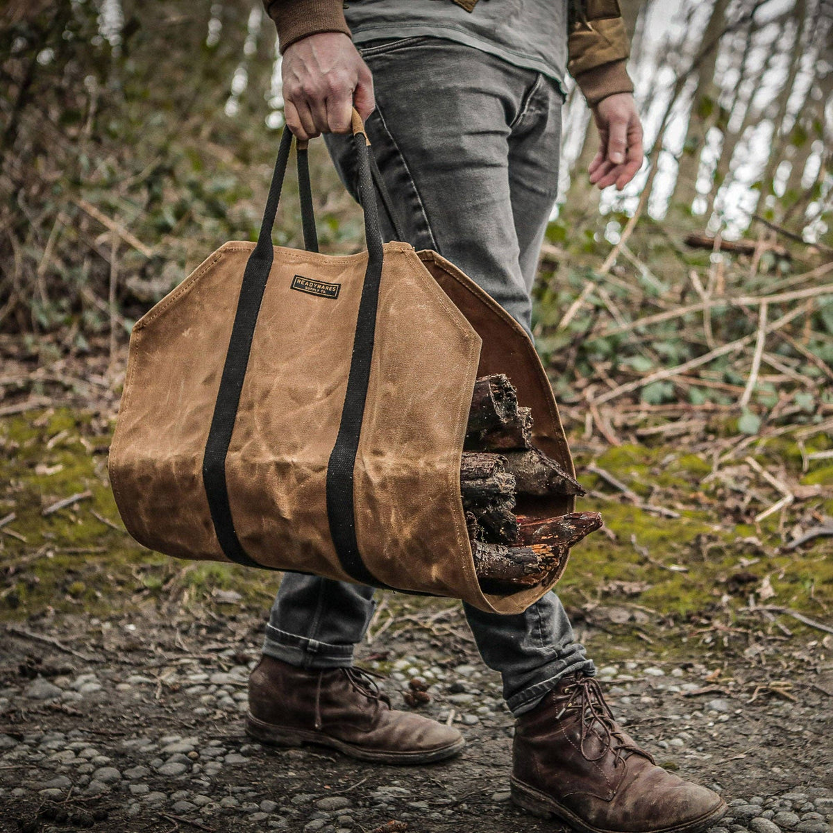 Person in jeans and boots carrying the tan waxed canvas firewood carrier full of logs through the woods