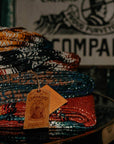Stack of folded striped Compadre blankets with a hang tag, in front of a Compadre sign