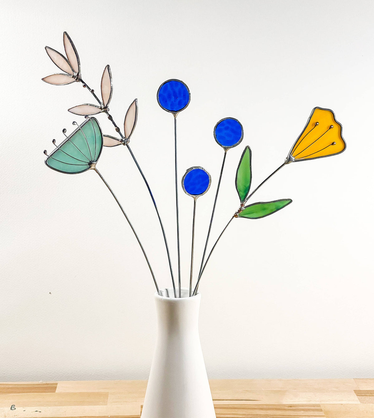 Stained glass bouquet of a yellow tulip, blue round flowers, aqua flower and sprig in a white vase