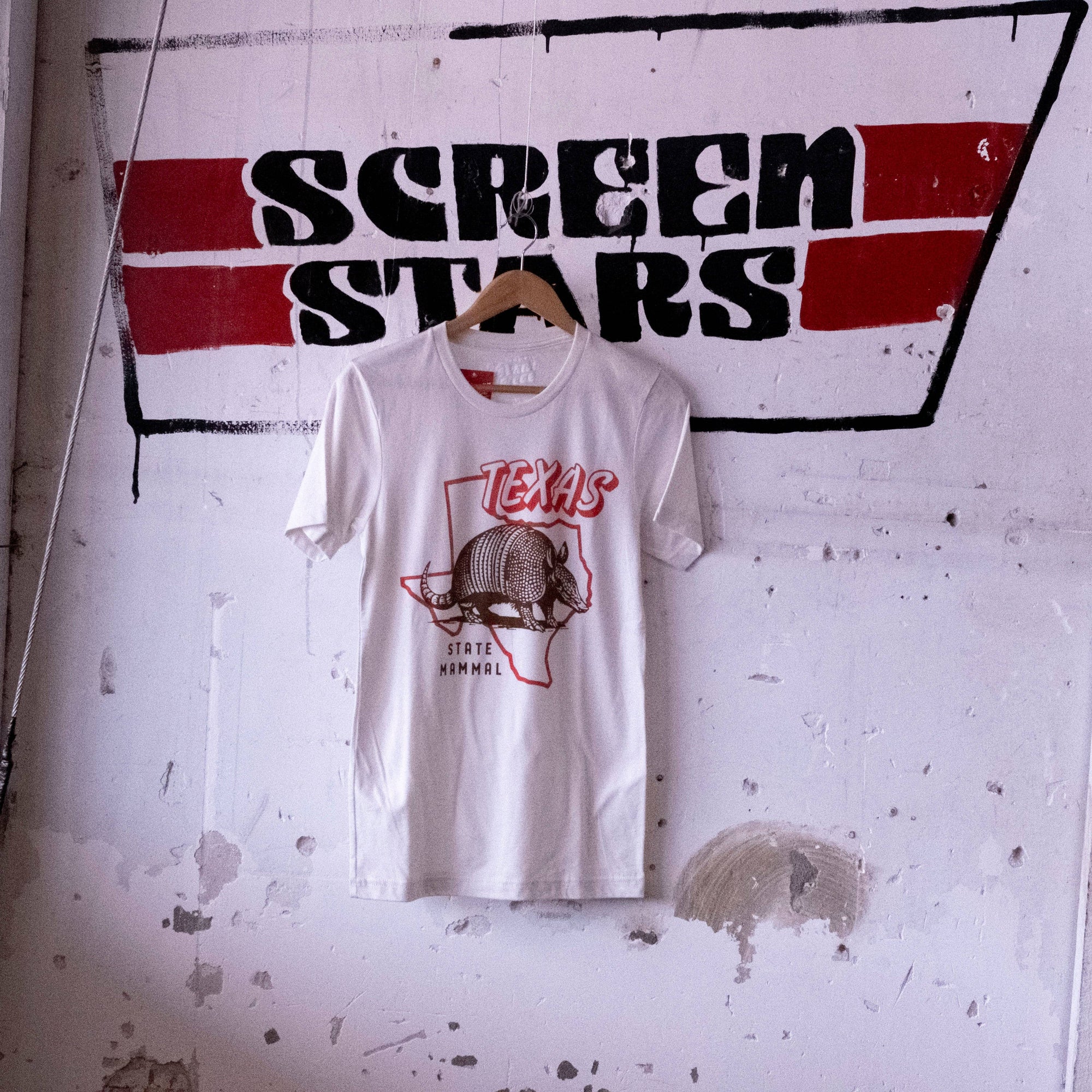 A white short-sleeve graphic T-shirt hanging on a wooden hanger against a distressed white wall. The shirt features a red and brown graphic of the state of Texas with an armadillo and the text 'TEXAS' and 'STATE CAPITAL'.