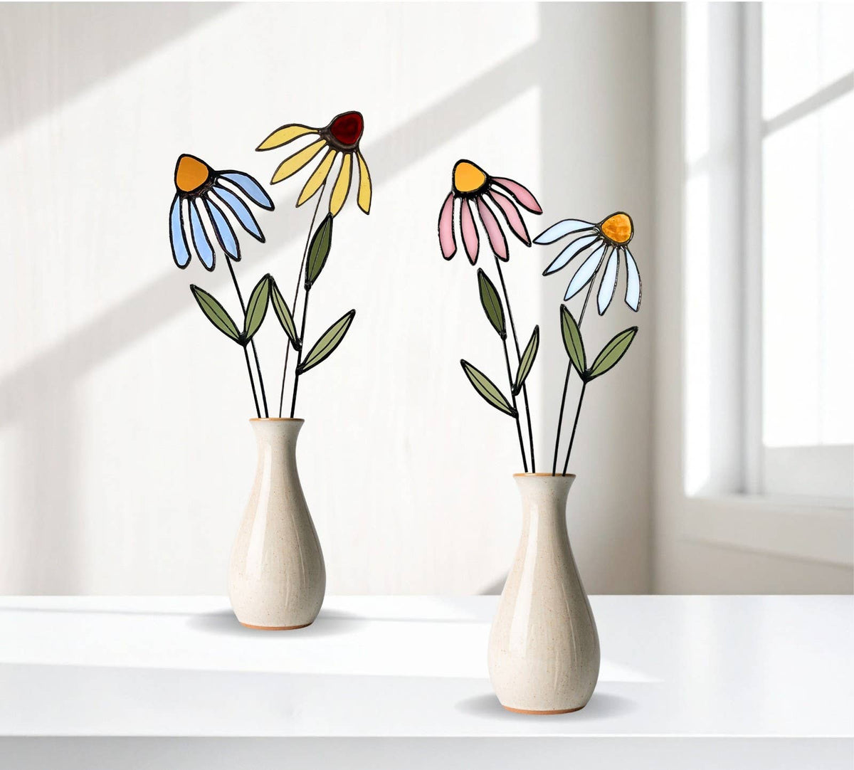 Two vases of stained glass flowers, including blue, yellow and pink coneflowers, on a white ledge