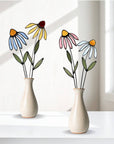 Two vases of stained glass flowers, including blue, yellow and pink coneflowers, on a white ledge