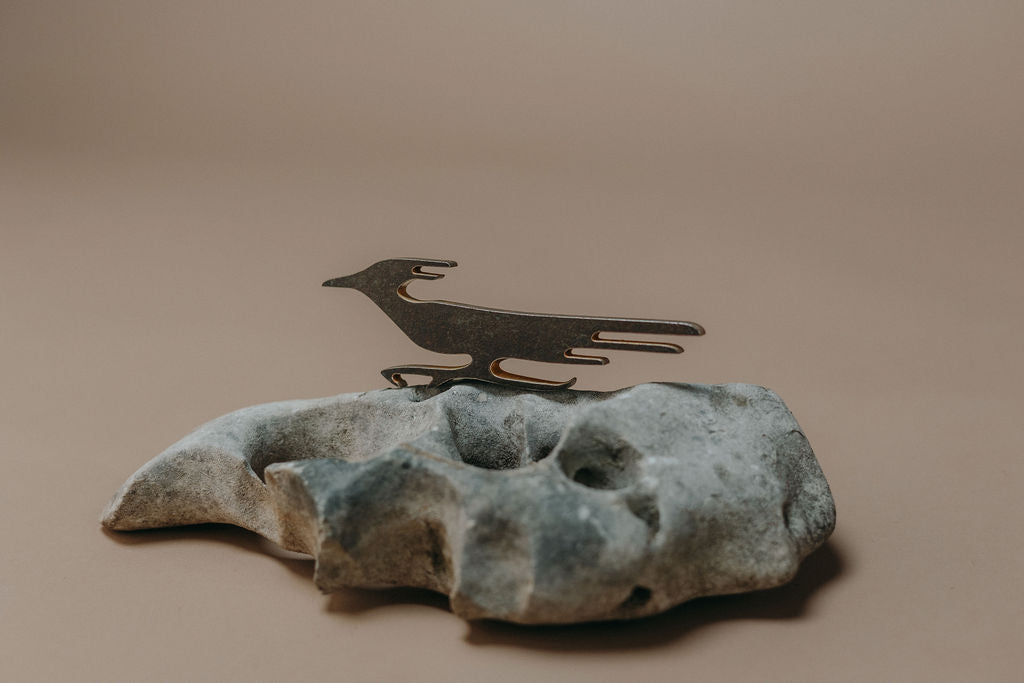 A dark metal bird silhouette candle holder mounted on a textured gray stone base, photographed against a solid warm beige background.