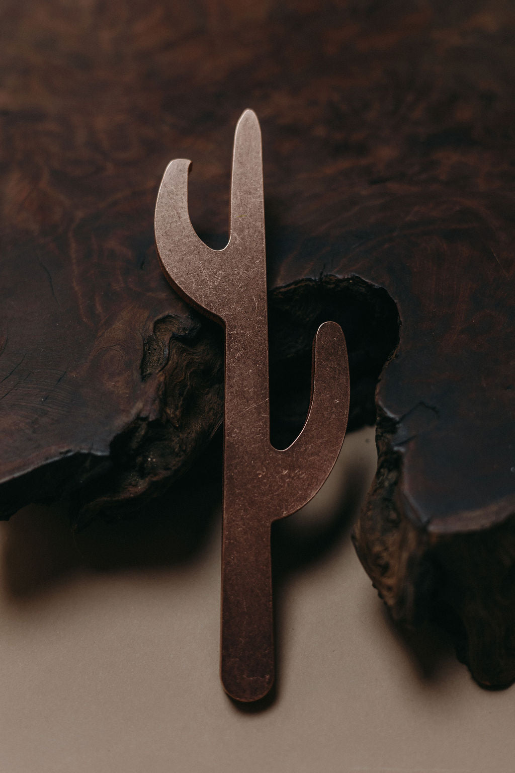 A metal cactus-shaped copper saguaro shaped bottle opener displayed against a dark wood bark background and neutral surface.

