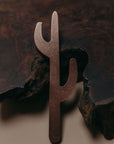 A metal cactus-shaped copper saguaro shaped bottle opener displayed against a dark wood bark background and neutral surface.

