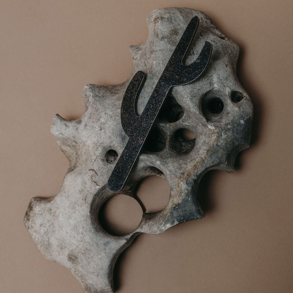 A black metal cactus bottle opener mounted on a textured, organic-shaped gray stone base, set against a solid warm brown background.