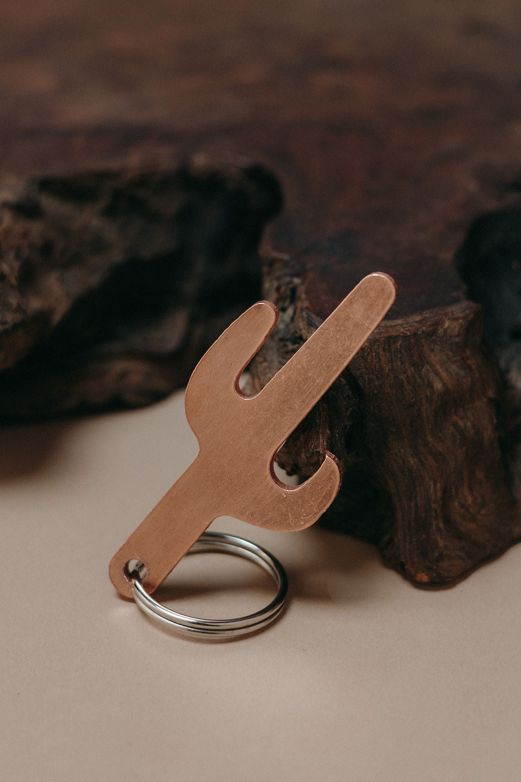 A cactus-shaped metal keychain tool with a bottle opener cutout and a silver key ring, photographed on a beige surface with dark wood in the background.