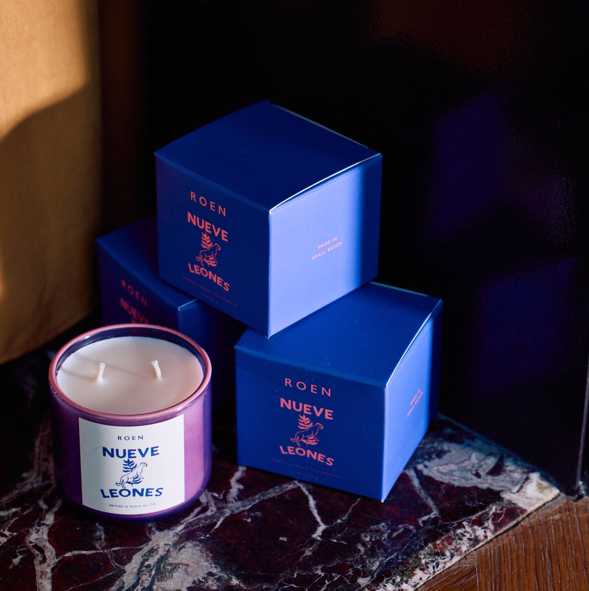 Nueve Leones candle in a painted jar next to stacked blue ROEN boxes on marble