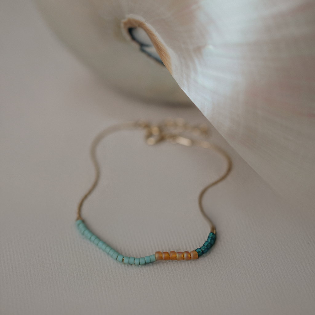A delicate gold chain bracelet with small turquoise and amber beads, resting on a textured light beige surface next to a white shell.