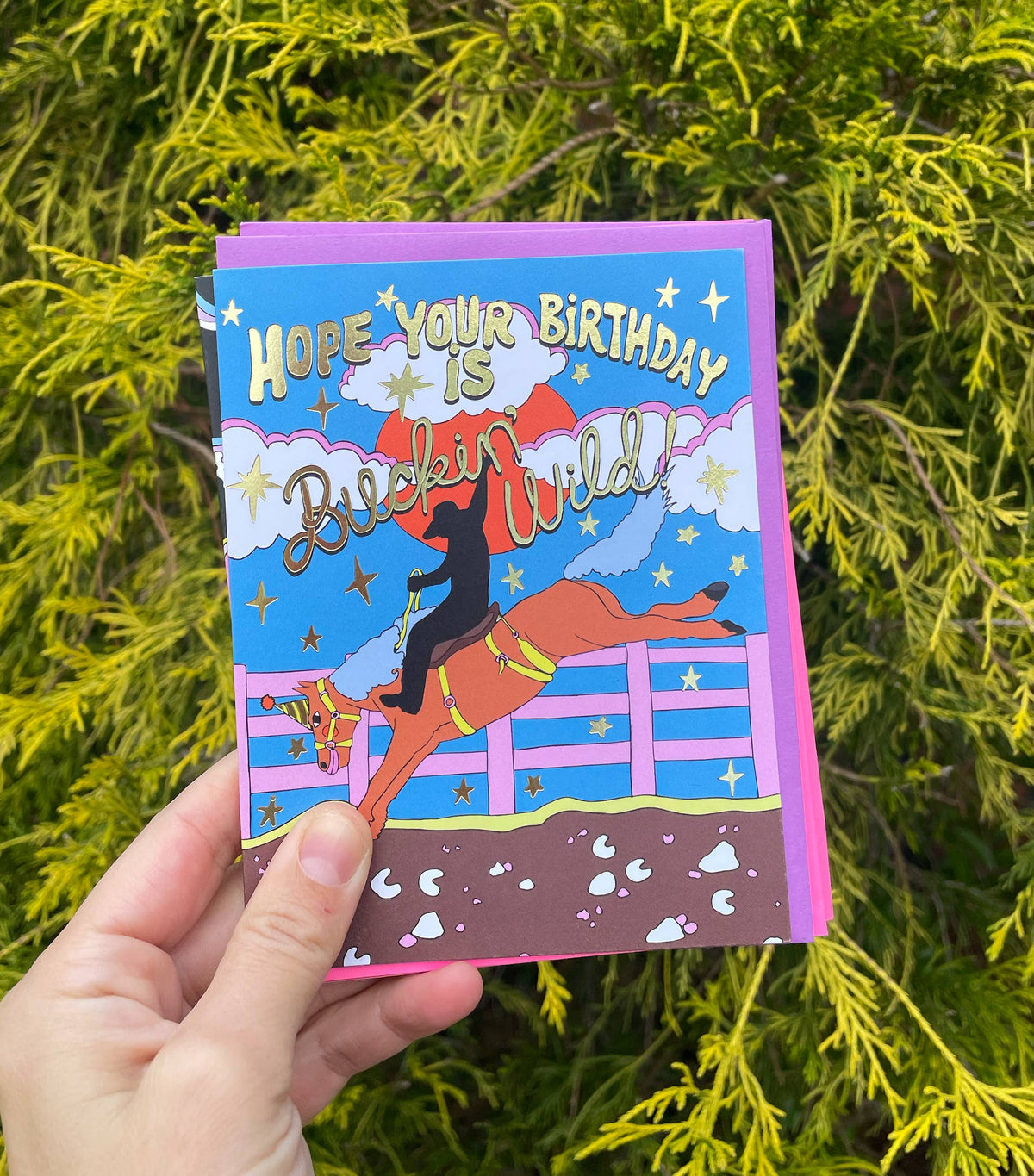 Hand holding a colorful birthday card with a horse and rider design against a green leafy background.