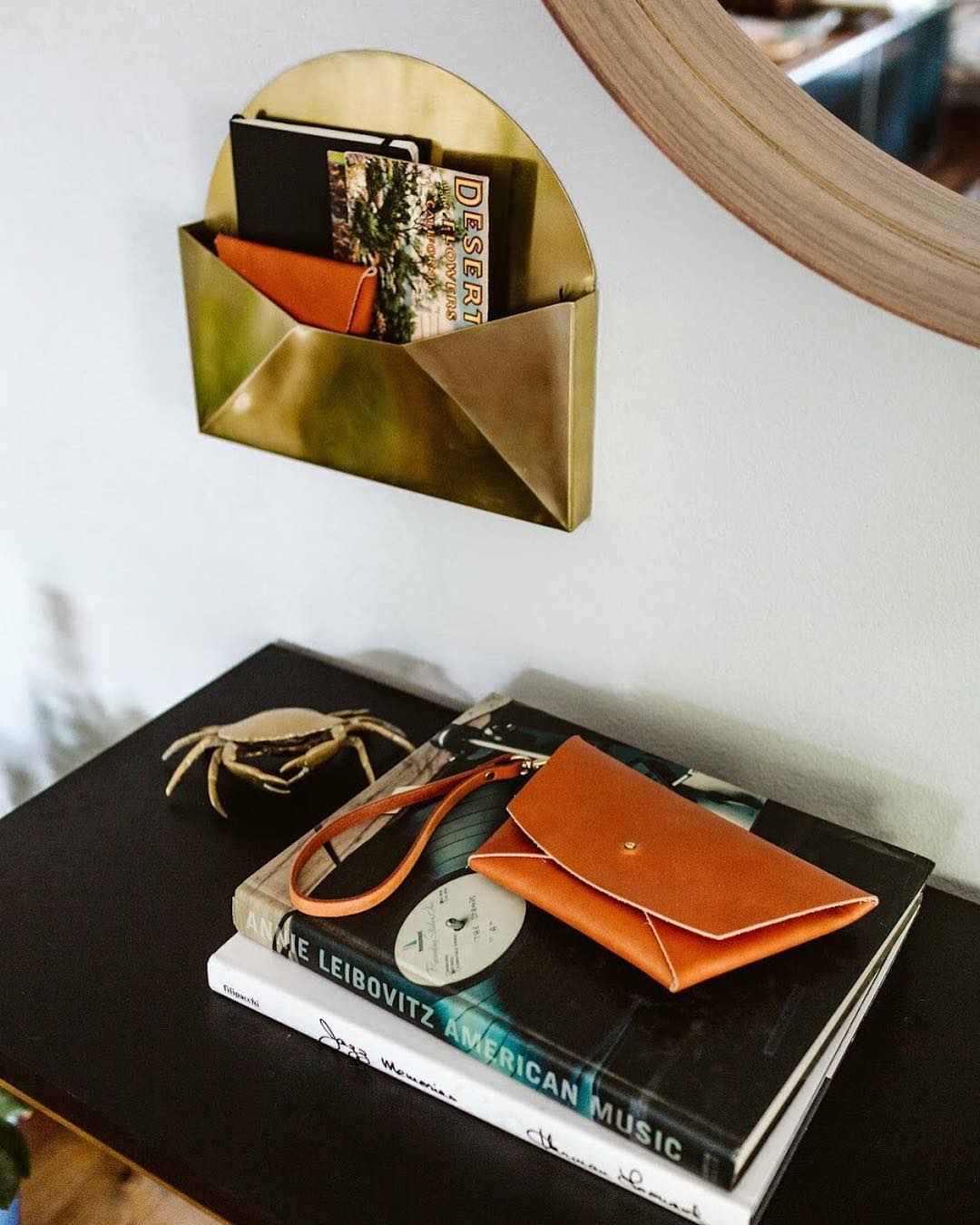 Tan leather clutch with strap on a stack of books on a dresser, brass wall pocket above