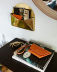 Tan leather clutch with strap on a stack of books on a dresser, brass wall pocket above