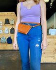 Model in a lavender tank and jeans wearing a tan leather clutch as a hip pack in the shop