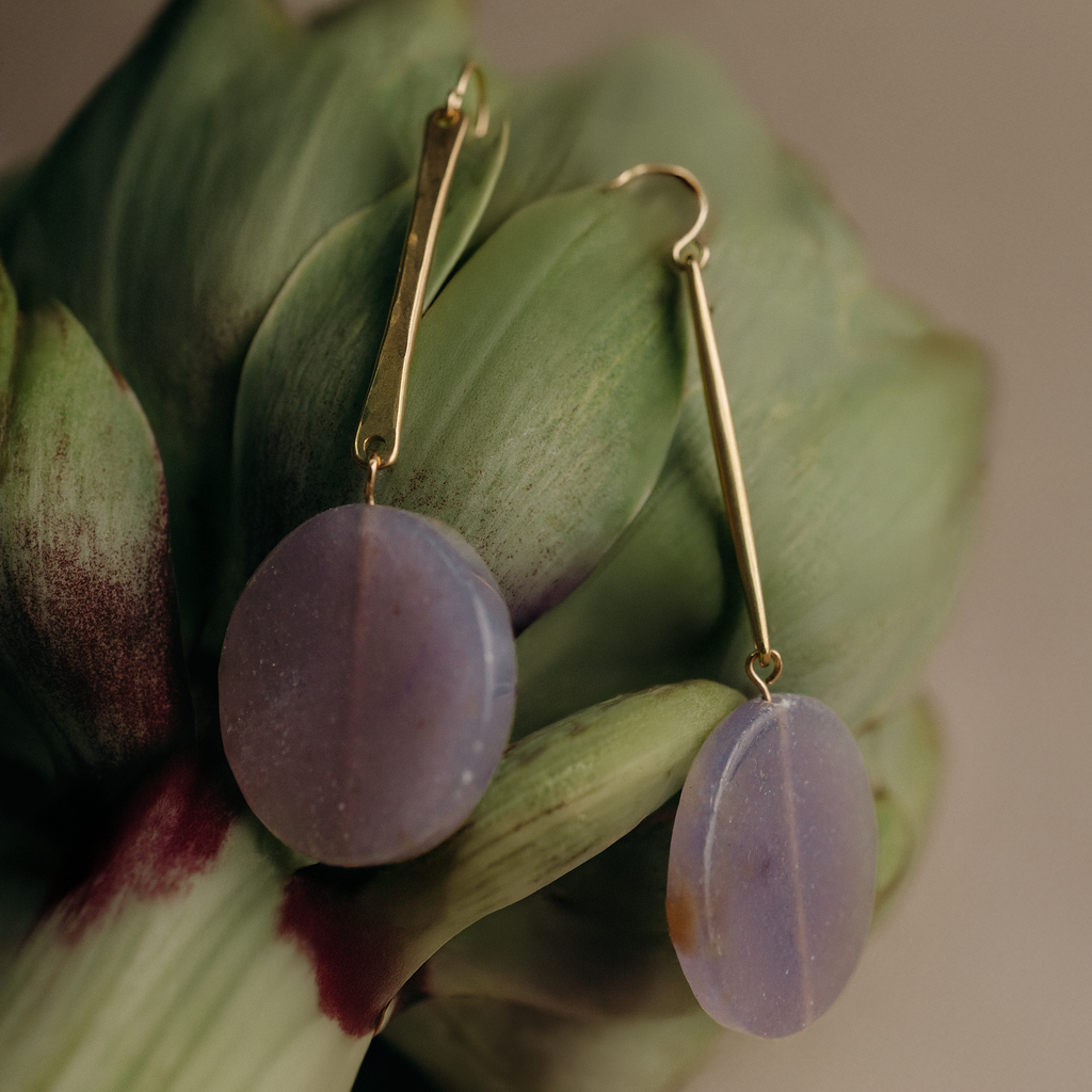A pair of gold drop earrings with oval lavender-purple stone pendants, displayed on the green petals of an artichoke against a soft beige background.