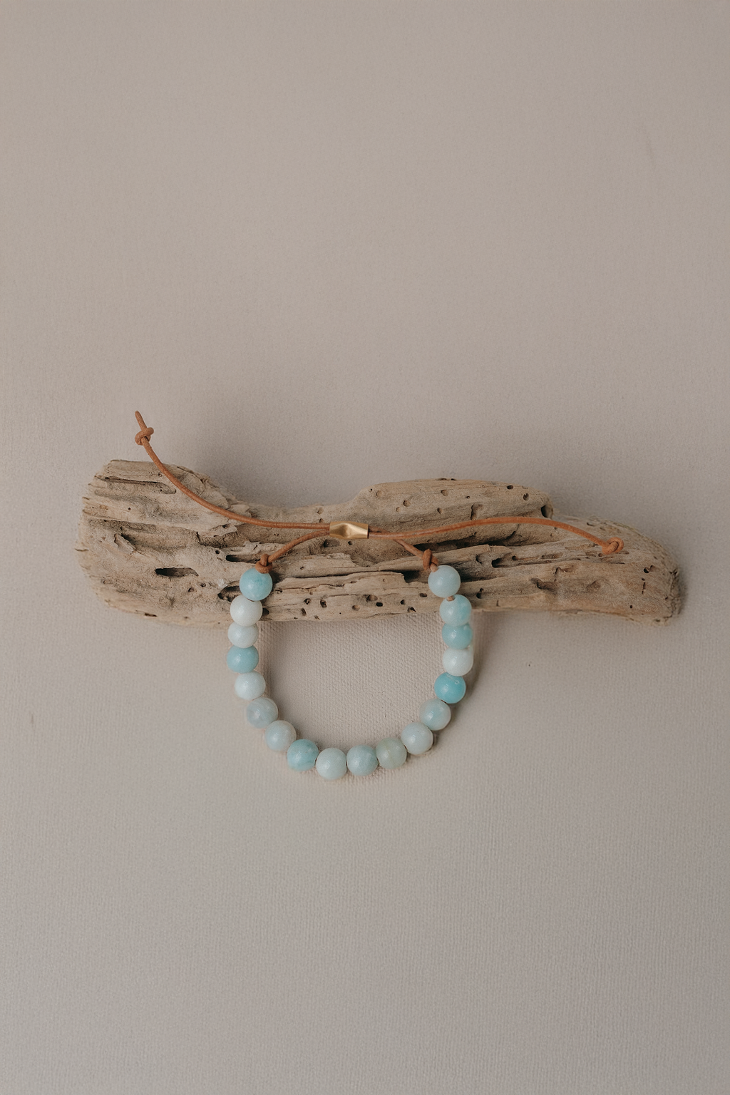 A beaded bracelet made of light blue and white round beads, draped over a piece of natural driftwood against a plain beige wall.