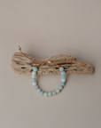 A beaded bracelet made of light blue and white round beads, draped over a piece of natural driftwood against a plain beige wall.