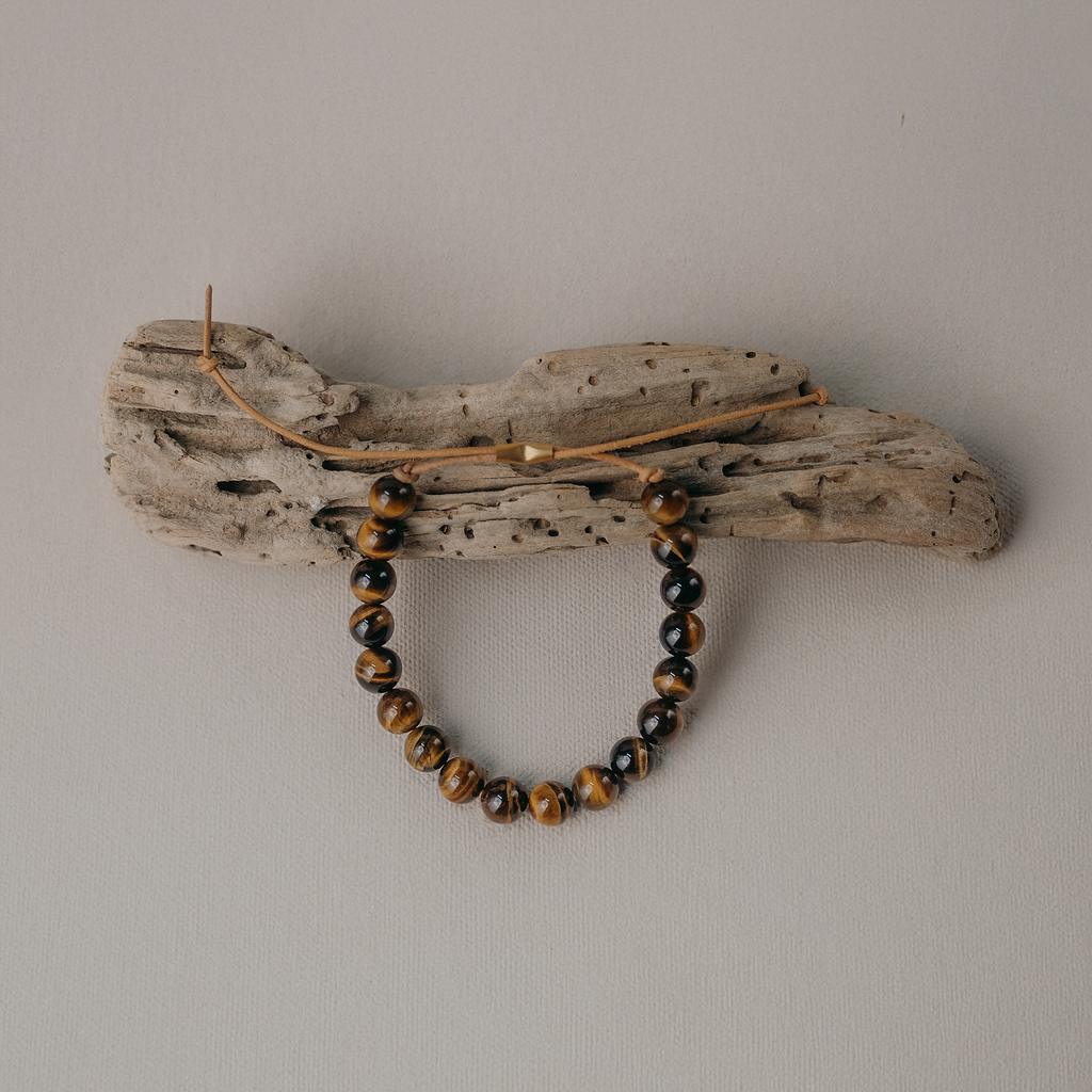 A beaded bracelet with brown and black marbled beads hangs from a rustic driftwood wall hook against a plain light gray wall.