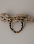 A beaded bracelet with brown and black marbled beads hangs from a rustic driftwood wall hook against a plain light gray wall.