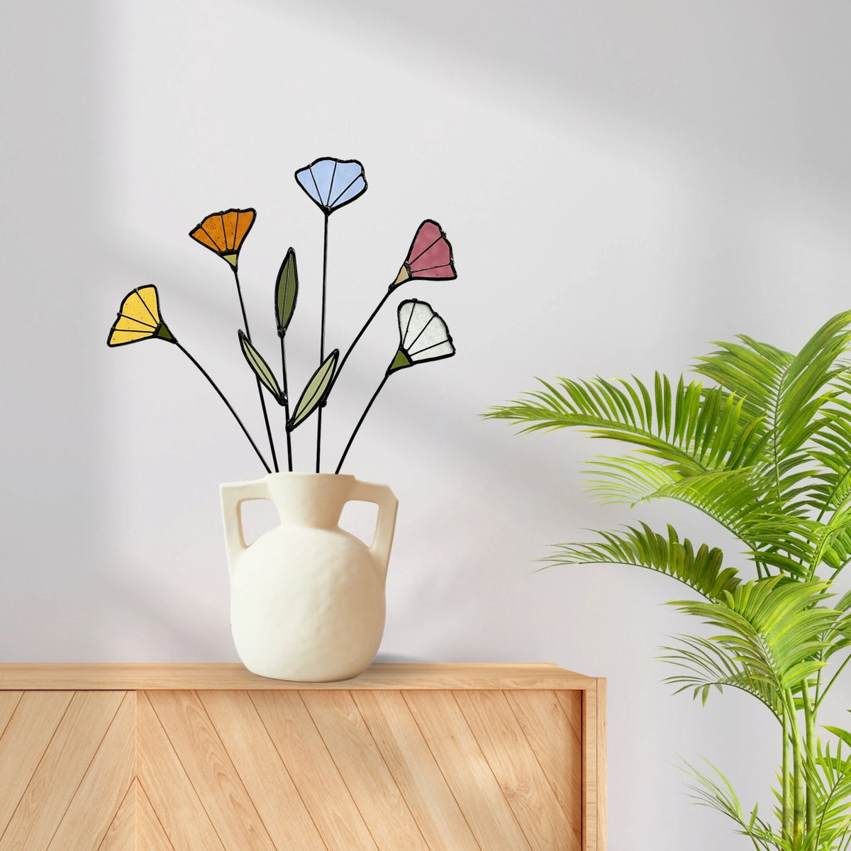 Stained glass wildflowers in a white handled vase on a wooden cabinet beside a palm