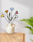 Stained glass wildflowers in a white handled vase on a wooden cabinet beside a palm