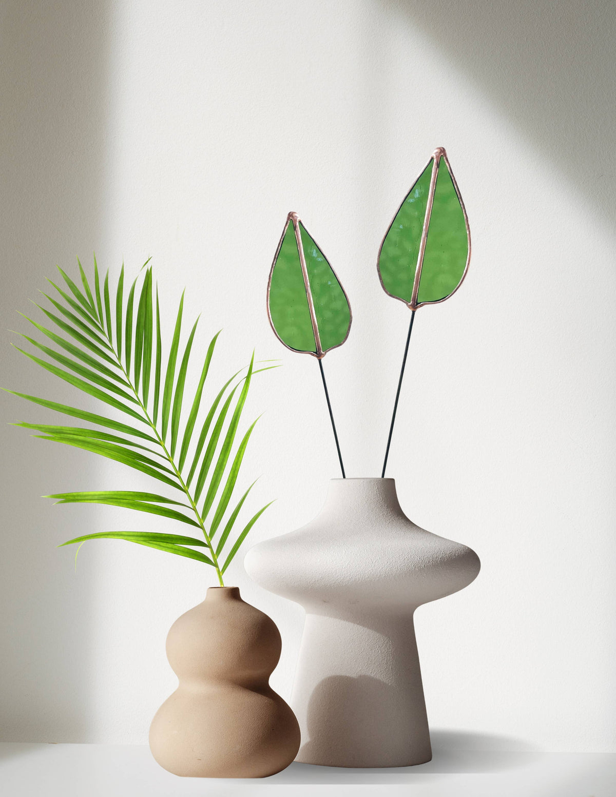Two green stained glass mulberry leaf stems in a white vase beside a tan vase with a palm frond